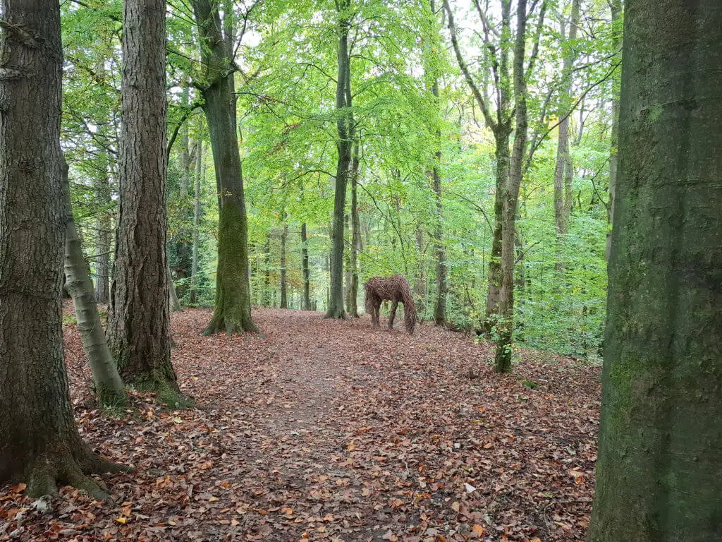A woodland path covered in fallen leaves with trees lining the sides. In the distance, a sculpture of a horse made from natural materials blends in.