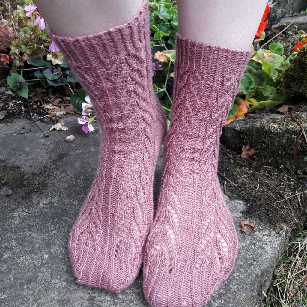 Soft pink knitted socks with intricate patterns on feet, worn by a person outdoors on stone steps. Surrounding greenery adds a vibrant touch.