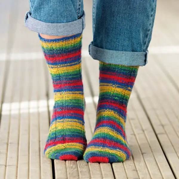 Close-up of feet wearing colourful, striped socks with rainbow hues, paired with rolled-up blue jeans on a light wooden floor, conveying a playful vibe. Winwick Mum yarn | West Yorkshire Spinners Signature 4ply