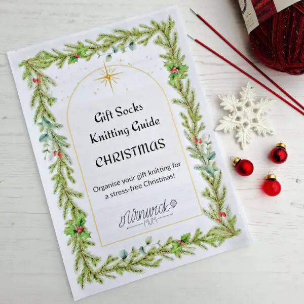 Winwick Mum gift knitting guide cover with Christmas theme, bordered by pine branches and holly. Festive decor includes baubles, a snowflake, a ball of yarn and knitting needles.