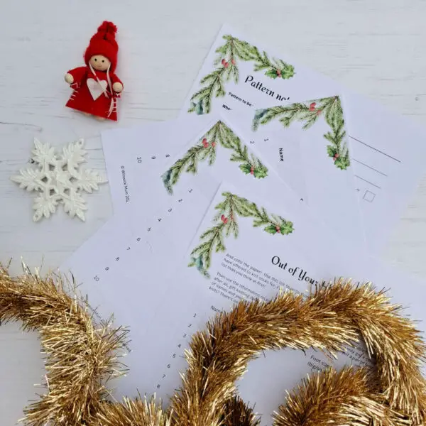 Festive scene with Christmas-themed worksheets featuring pine and holly designs, a small snowflake ornament, a red-clad doll, and gold tinsel on a wooden surface.