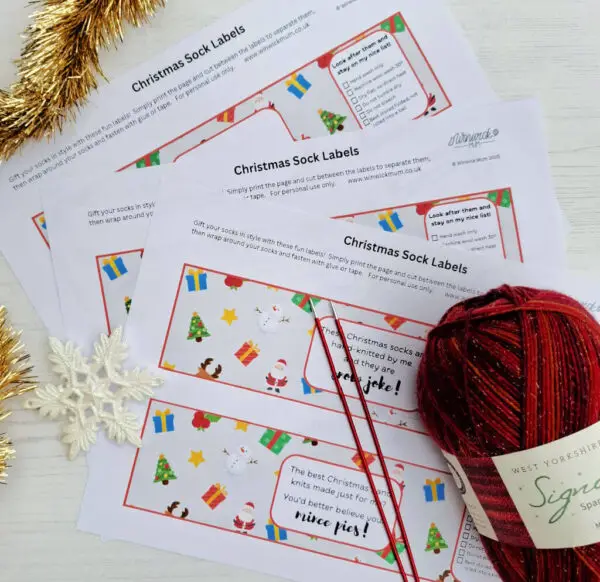 Sheets of Christmas sock labels featuring festive icons like snowmen and gifts are placed on a table. Nearby are red knitting needles, a sparkly red yarn ball, and gold tinsel. The scene feels cosy and festive.