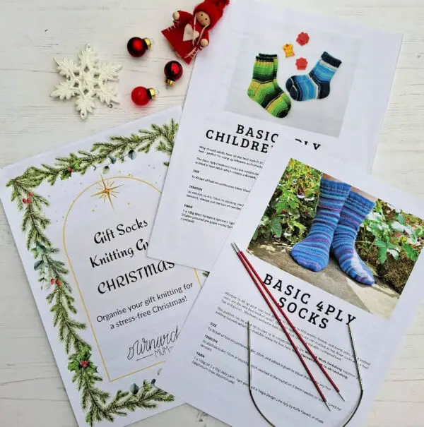 Two knitting patterns and a gift knitting guide are laid out on a white surface. The guide features a Christmas theme, the patterns are children's socks, and adult socks. Nearby are knitting needles, a crocheted snowflake, and festive decorations, including red baubles and a small doll.