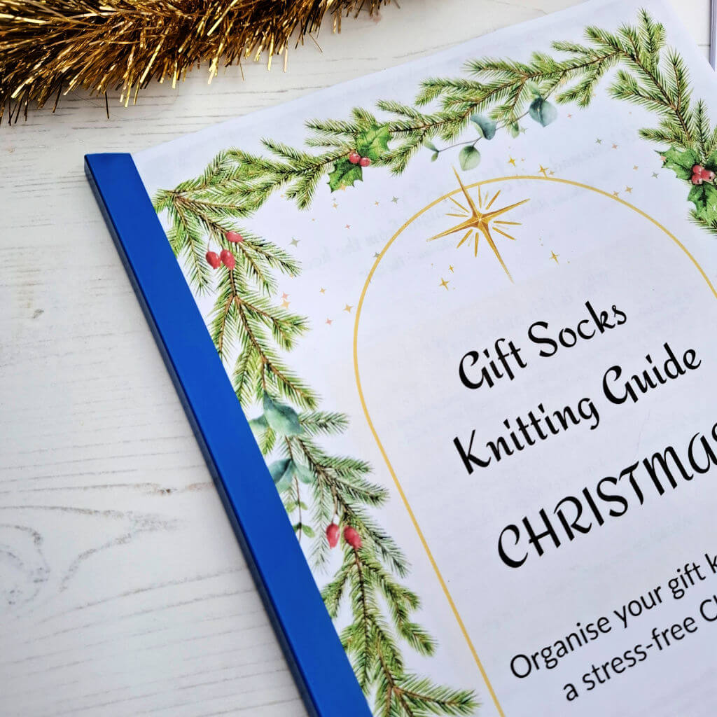 A close up of the blue spine added to a printed festive knitting guide which lies on a white wood surface. It features a Christmas pine and holly border with a golden star accent.