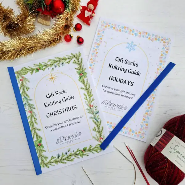 Two Winwick Mum knitting guides titled 'Gift Socks Knitting Guide: Christmas' and 'Holidays' with festive designs and needles on a white surface. One has a blue spine fitted and the second spine lies across one of the guides