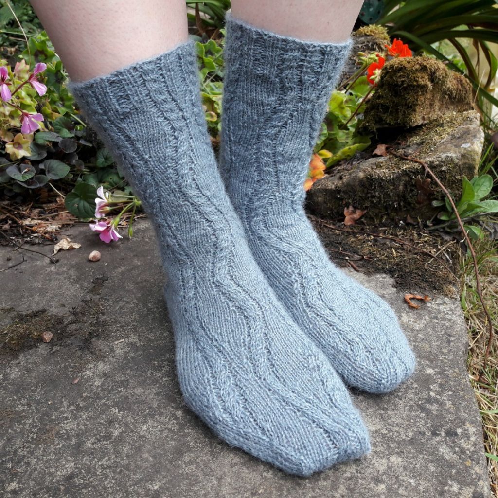 Close-up of feet in light blue knitted socks with a cable pattern, standing on a stone path. Colourful flowers and green foliage surround the scene.