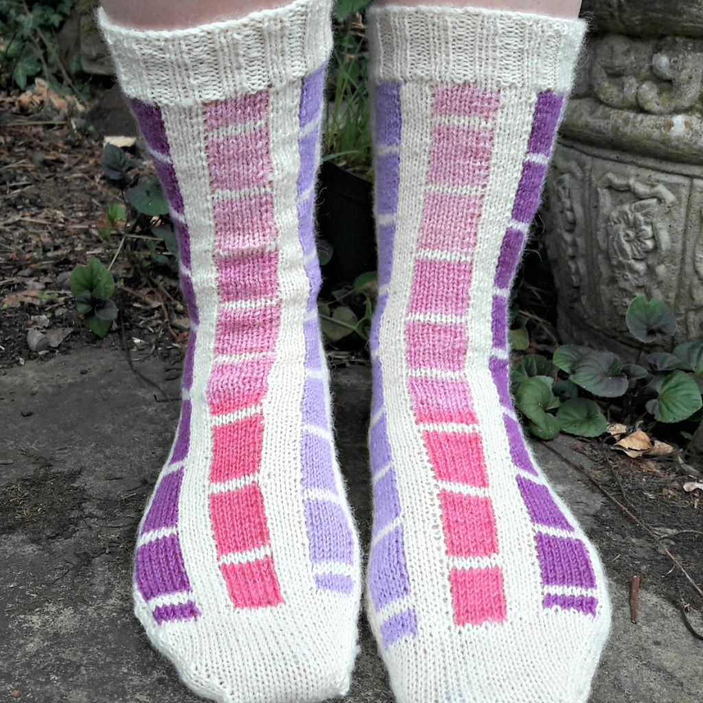Close-up of two feet in colourful knitted socks with a grid pattern. The socks feature pink, purple, and white squares. They rest on a stone path surrounded by greenery.