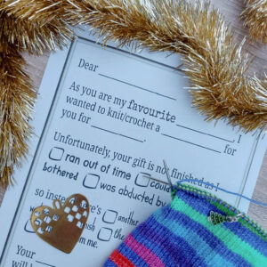 Knitting-themed card with humorous message about unfinished gifts surrounded by golden tinsel. A colourful knitted piece and heart-shaped cutout are nearby.