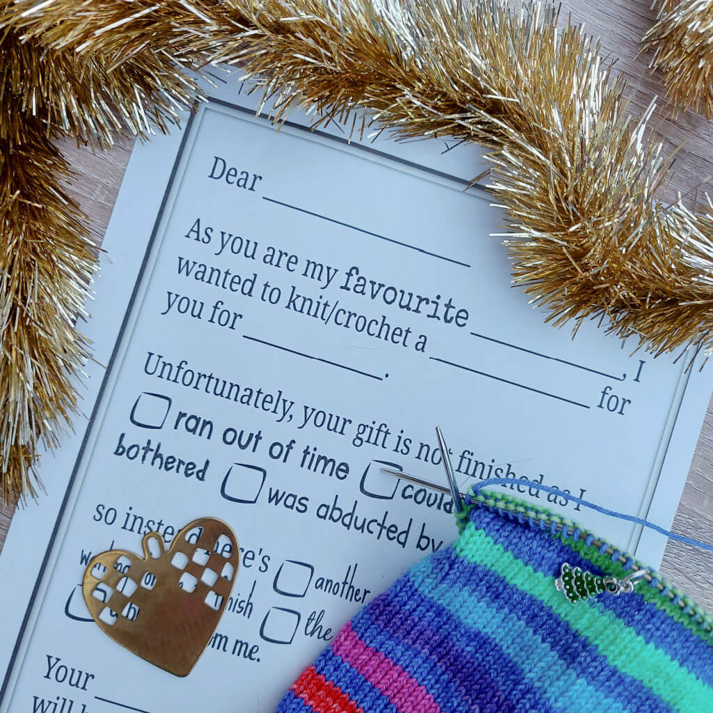 Knitting-themed letter with humorous message about unfinished gifts surrounded by golden tinsel. A colourful knitted piece and heart-shaped cutout are nearby.