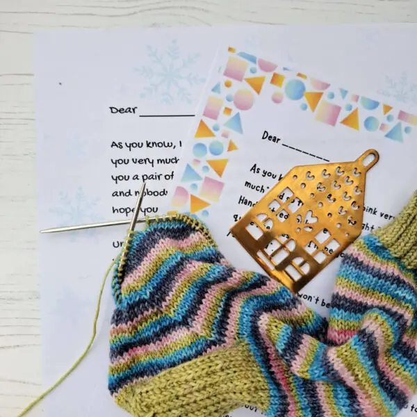 Knitting in progress with multicoloured yarn and needles atop festive letters. A gold decorative stitch marker lies nearby. Cosy and creative ambiance.