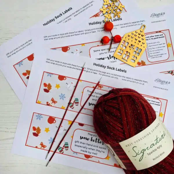 Holiday sock labels with festive designs are spread out on a table. Festive knitting supplies, including red yarn and needles, create a cosy, seasonal atmosphere.