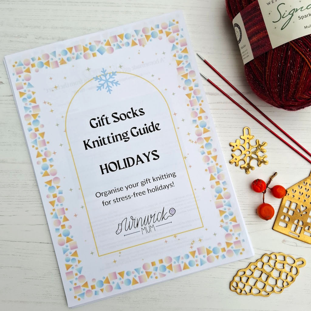 Winwick Mum gift knitting guide cover with Christmas theme, bordered by pine branches and holly. Festive decor includes a brass ornaments, a ball of yarn and knitting needles.