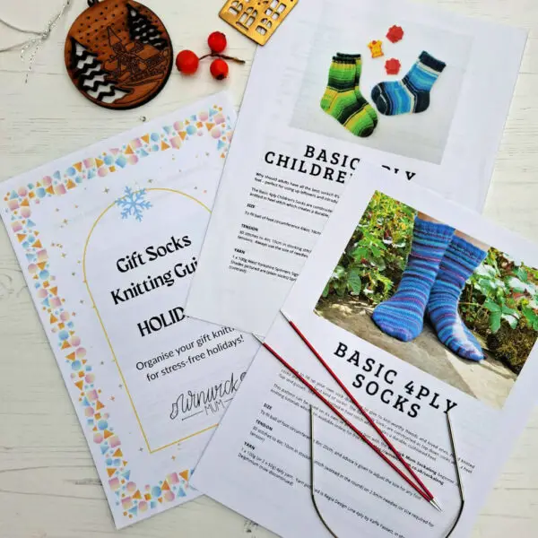 Holiday gift guide for knitting socks, plus children's and adult sock patterns, are spread on a table. A festive design and knitting needles are visible, creating a cosy, creative atmosphere.