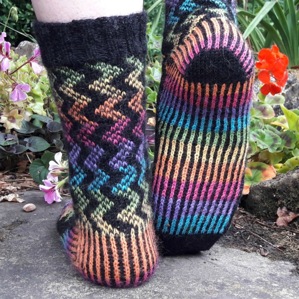 Close-up of two feet wearing vibrant, hand-knitted socks. One sock has wavy zigzag patterns; the other features vertical stripes. Background includes colourful flowers and greenery, creating a lively and cosy atmosphere.