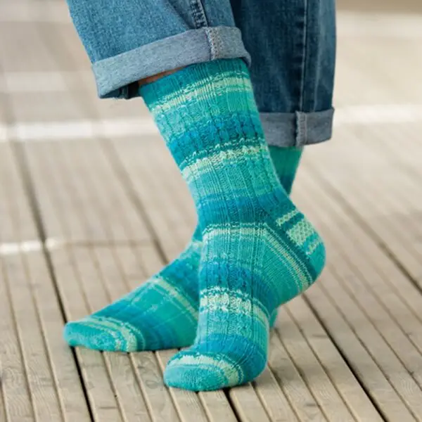 Person wearing vibrant teal patterned socks and cuffed blue jeans, standing on a wooden deck, conveying a cosy and relaxed vibe. Winwick Mum yarn | West Yorkshire Spinners Signature 4ply