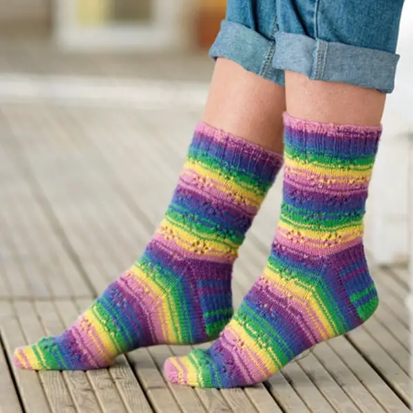 Person wearing colourful, striped wool socks featuring purple, green, yellow, and blue hues. The background shows a light wooden floor, conveying warmth. Winwick Mum yarn | West Yorkshire Spinners Signature 4ply