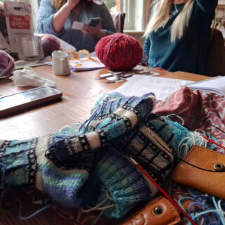 A cosy knitting group gathers around a wooden table covered with colourful yarn, knitting tools, and notes. People are engaged, creating a warm, collaborative atmosphere.