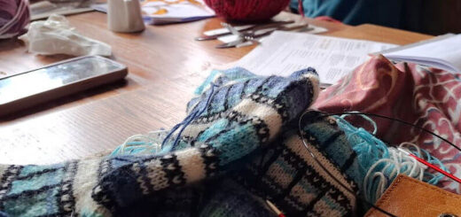 A cosy knitting group gathers around a wooden table covered with colourful yarn, knitting tools, and notes. People are engaged, creating a warm, collaborative atmosphere.
