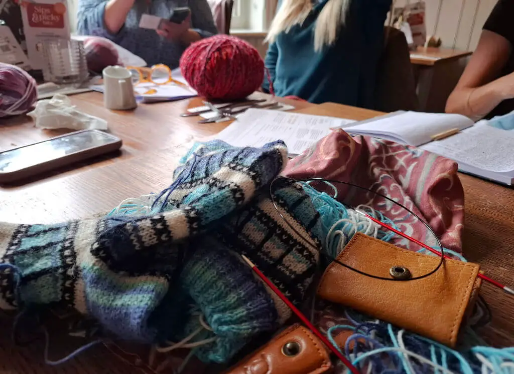 A cosy knitting group gathers around a wooden table covered with colourful yarn, knitting tools, and notes. People are engaged, creating a warm, collaborative atmosphere.
