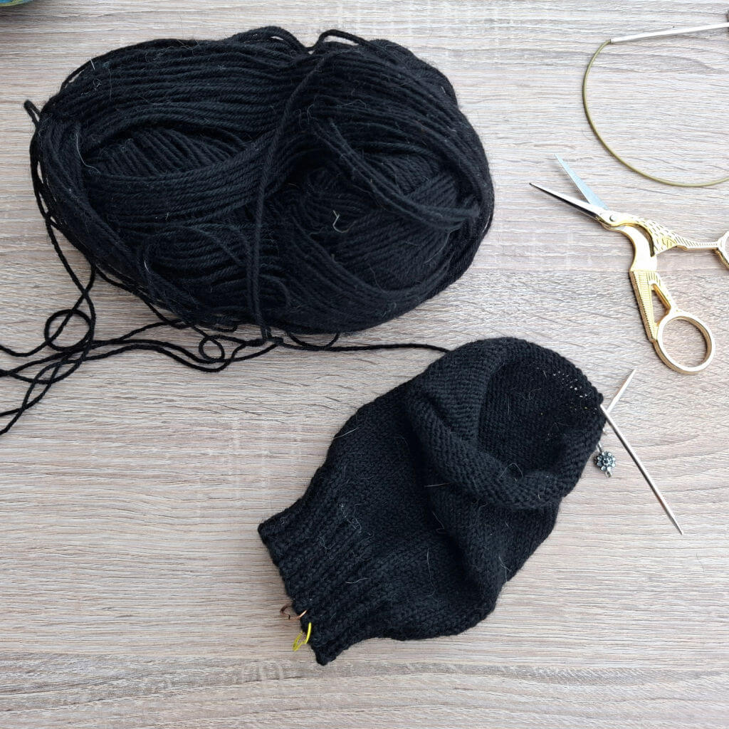 Black yarn and a partially knitted sock lie on a wooden surface. Nearby are golden embroidery scissors and a circular knitting needle. Cosy, creative tone.