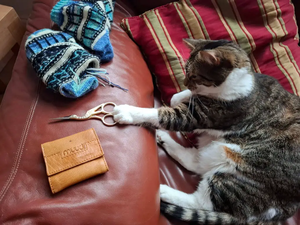 A tabby cat lies on a red couch with a paw on ornate scissors. Nearby are knitted blue socks and a tan leather needle case. The scene is cosy and playful.