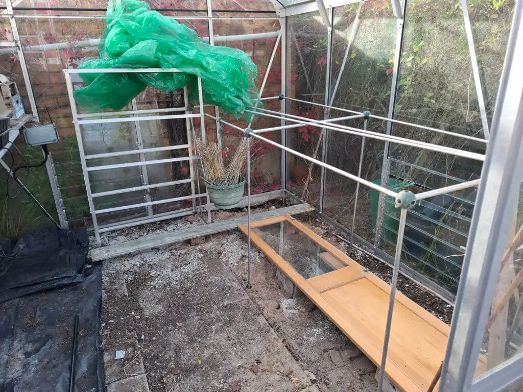 A small, empty greenhouse with metal frames and a wooden bench inside.