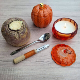 A wooden table displays a swede, spoon, peeler, orange pumpkin jar, and burning candle in a pumpkin-shaped holder next to the lid, creating a cosy autumn atmosphere.
