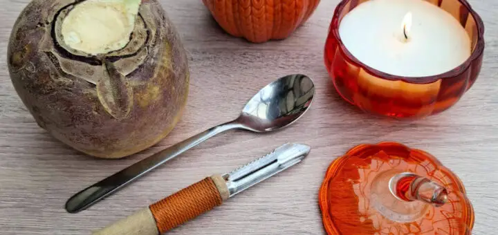 A wooden table displays a swede, spoon, peeler, orange pumpkin jar, and burning candle in a pumpkin-shaped holder next to the lid, creating a cosy autumn atmosphere.