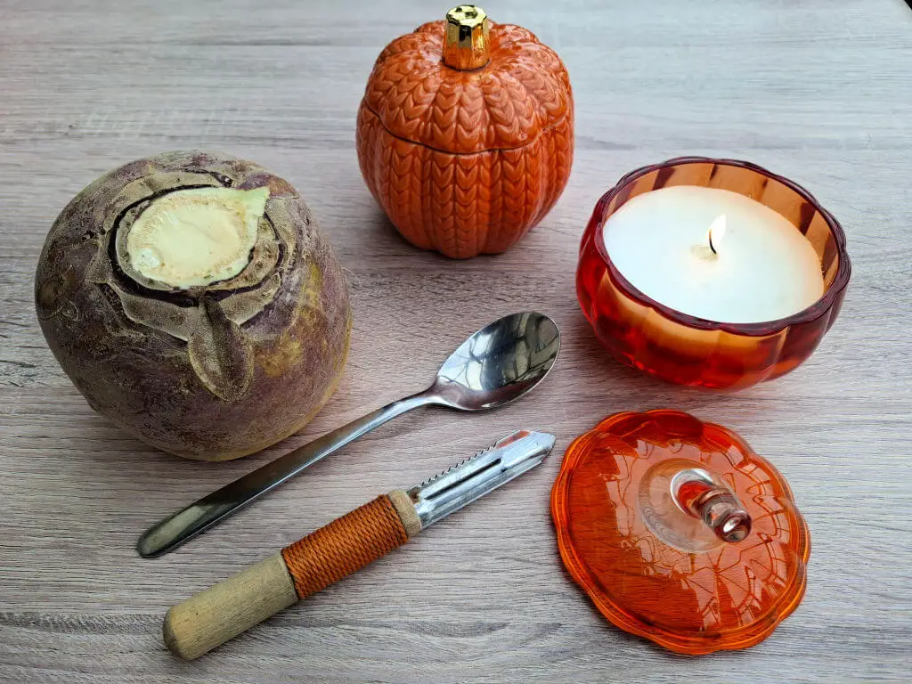 A wooden table displays a swede, spoon, peeler, orange pumpkin jar, and burning candle in a pumpkin-shaped holder next to the lid, creating a cosy autumn atmosphere.