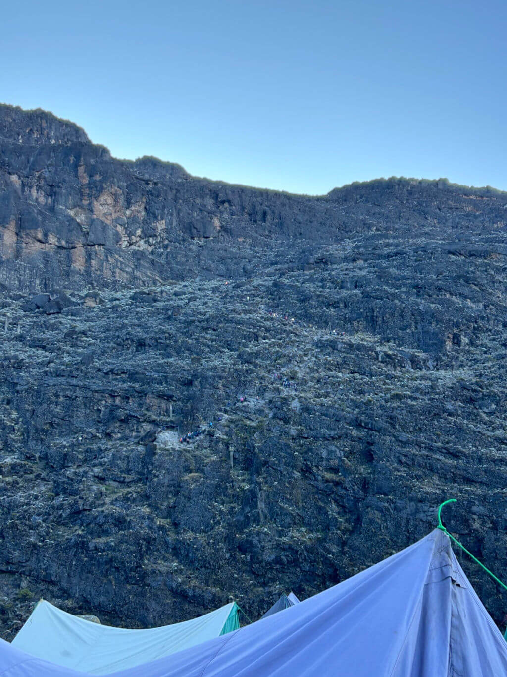 Rocky landscape of the Barranco Wall, Mount Kilimanjaro, under a clear blue sky, with tents visible at the bottom. The landscape conveys a sense of rugged beauty and tranquility.