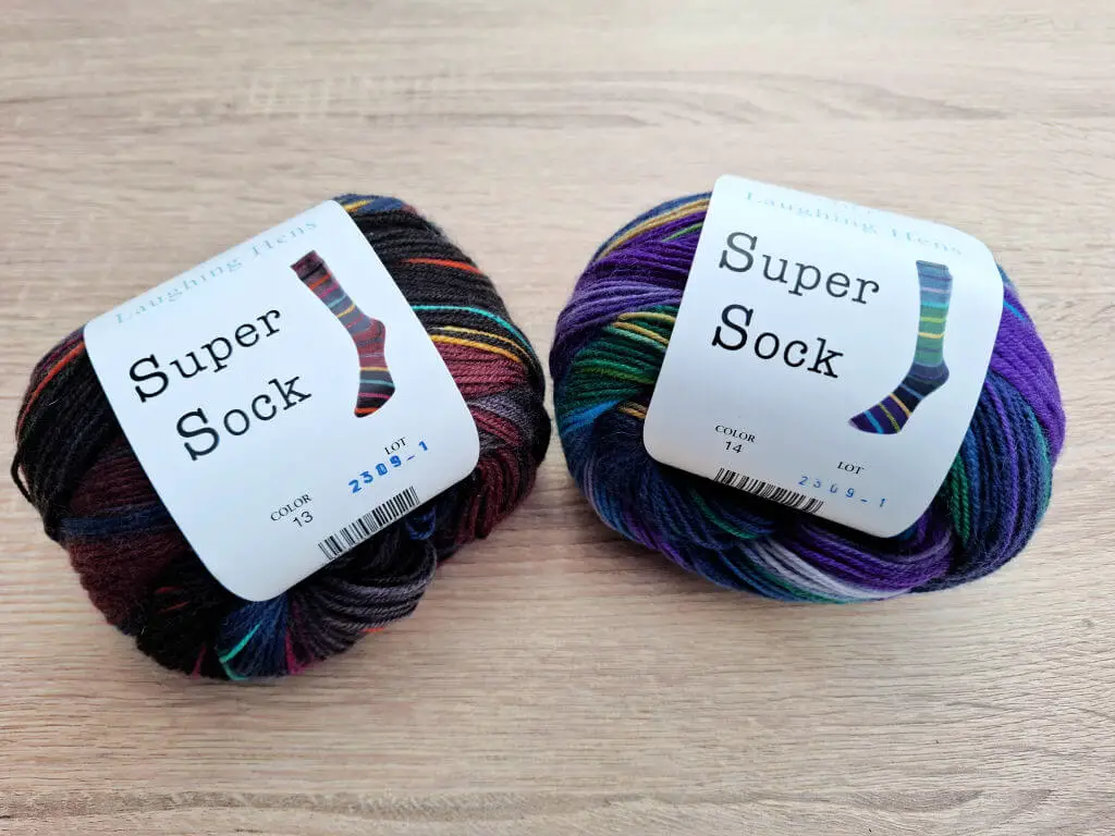 Two balls of multicoloured yarn labeled "Super Sock" on a light wooden surface. Colours include dark hues and bright tones, suggesting vibrant sock designs.