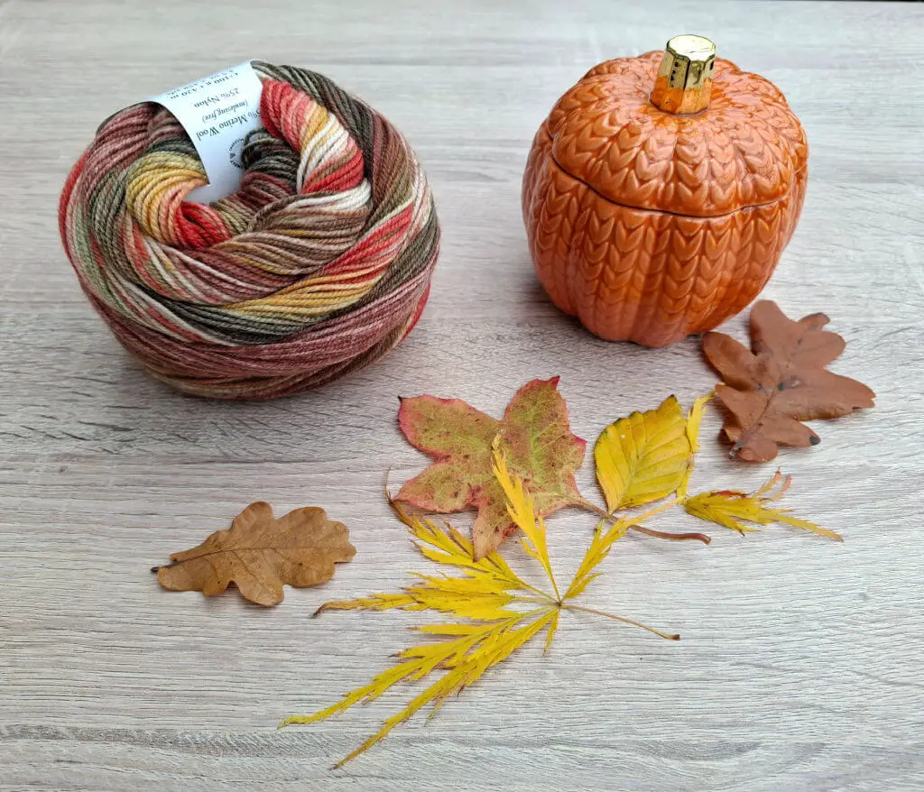 A ball of multicoloured yarn and a ceramic pumpkin sit on a wooden surface, surrounded by autumn leaves in shades of brown, red, and yellow. The scene conveys a cosy fall atmosphere.