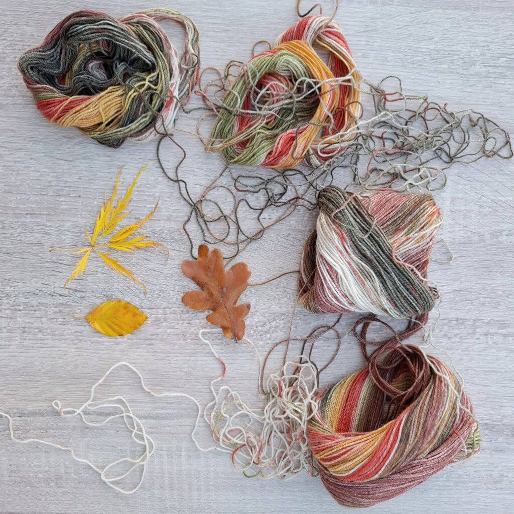 Four balls of multicoloured yarn with tones of red, green, and yellow are tangled on a wooden surface. Three autumn leaves add a touch of seasonal warmth.