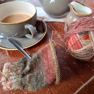 A cosy scene with a cup of tea, a teapot, and a jug on a wooden table. A partially knitted item and colourful yarn ball suggest a tranquil knitting session.