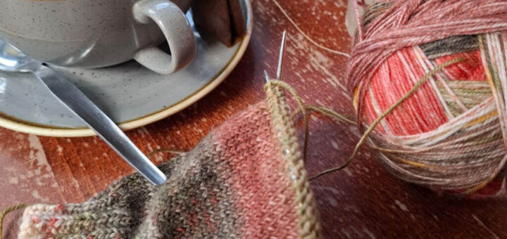 A cosy scene with a cup of tea, a teapot, and a jug on a wooden table. A partially knitted item and colourful yarn ball suggest a tranquil knitting session.