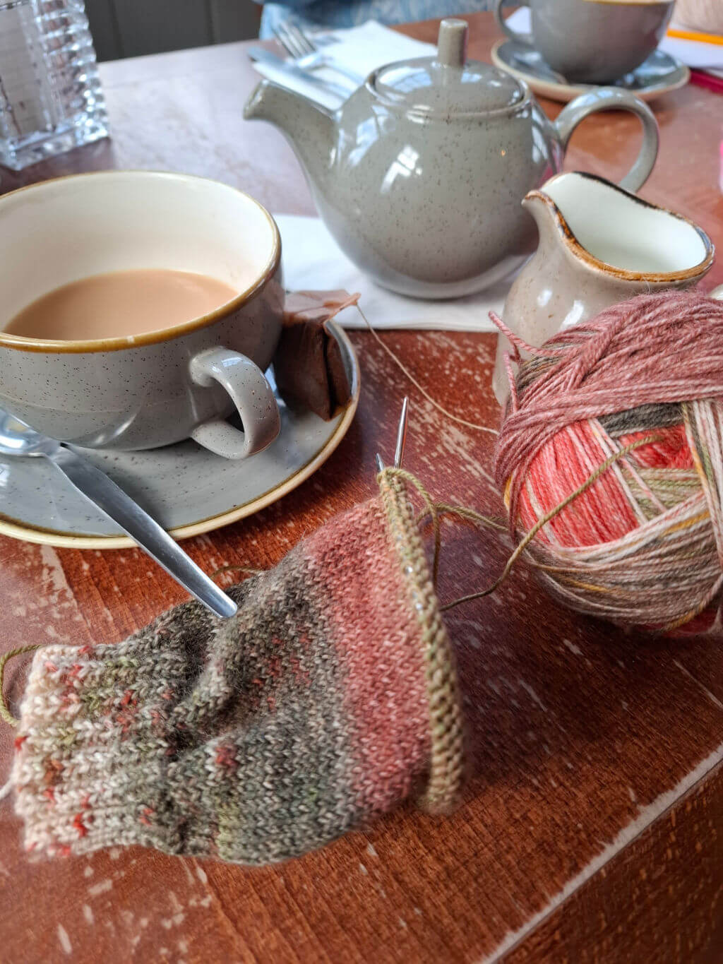 A cosy scene with a cup of tea, a teapot, and a jug on a wooden table. A partially knitted item and colourful yarn ball suggest a tranquil knitting session.