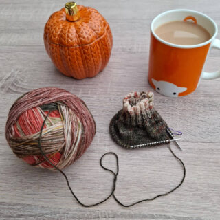 A cosy scene with a ball of multicoloured yarn partially knitted, a pumpkin-shaped container, and a mug of tea on a wooden table. Warm autumn vibes.