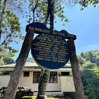Sign at Machame Gate, Kilimanjaro National Park, with route distances and times. Surrounded by trees, under a clear blue sky.