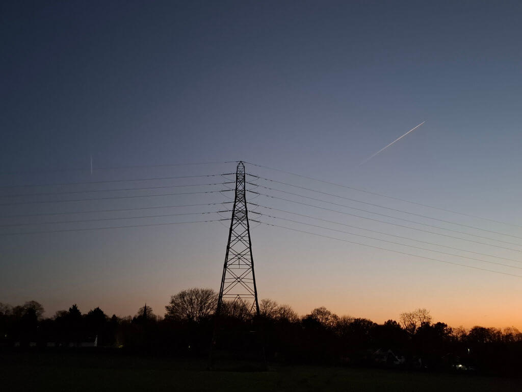 A silhouette of an electricity pylon stands against a dusk sky with a subtle gradient from deep blue to orange. A jet trail crosses the sky, above dark tree outlines.