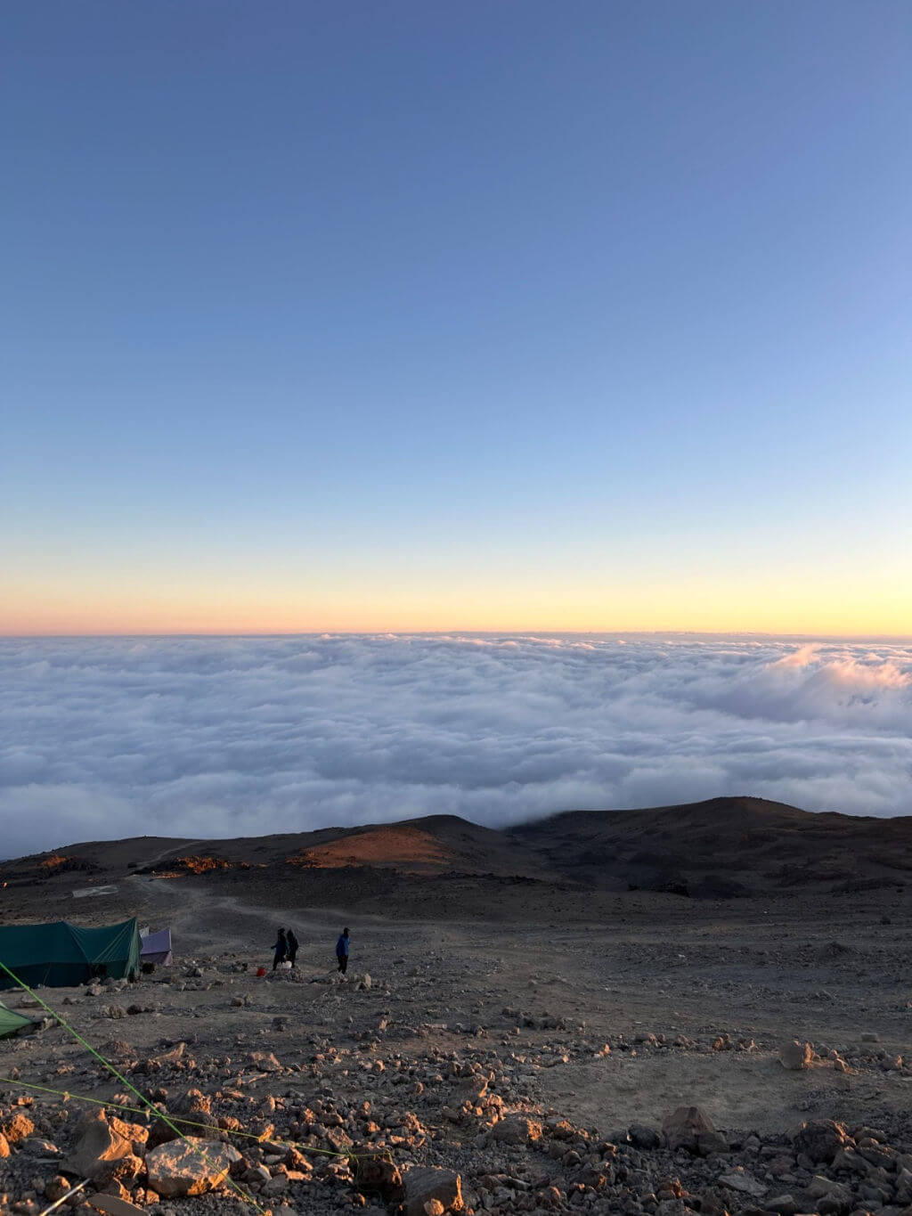 A rocky mountain landscape at one of the camps on Mount Kilimanjaro during sunset, with a vast sea of clouds below and a clear blue sky above. Two people and tents are visible in the foreground.