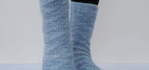 Two legs wearing light blue hand-knitted socks with a textured zigzag pattern, standing on a light wooden floor against a neutral background. Cosy and warm vibe.