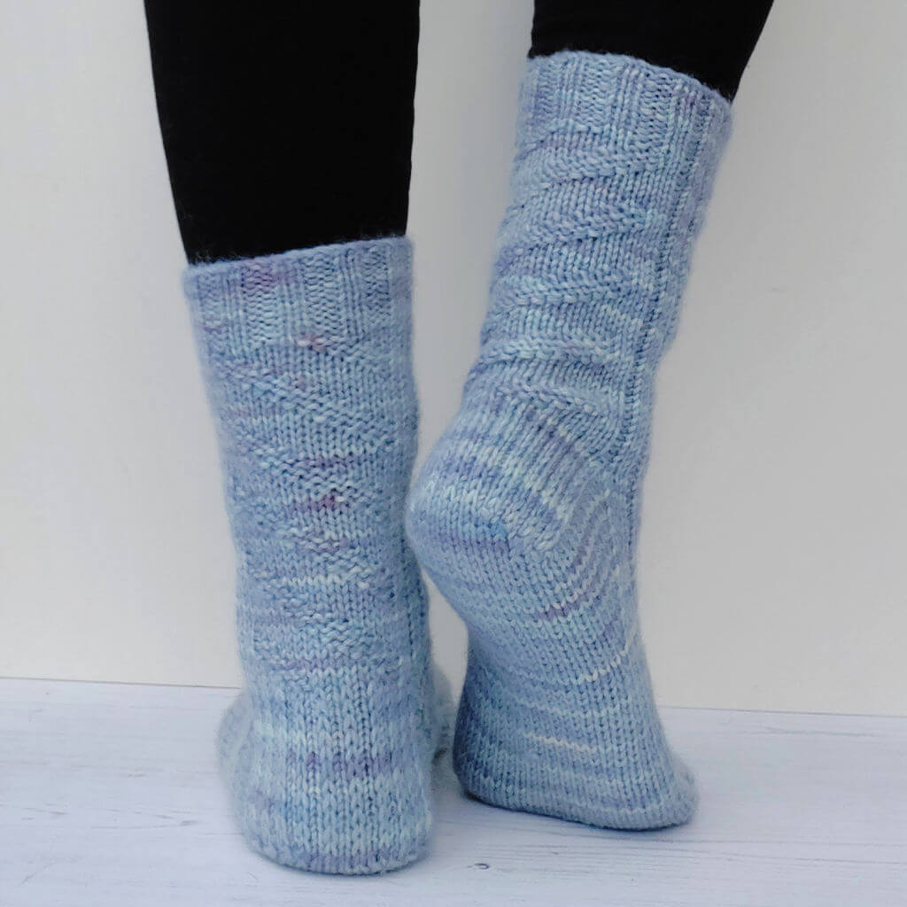 Close-up of two feet wearing soft, light blue knitted socks, standing on a white wooden floor. The texture appears cosy and slightly textured.