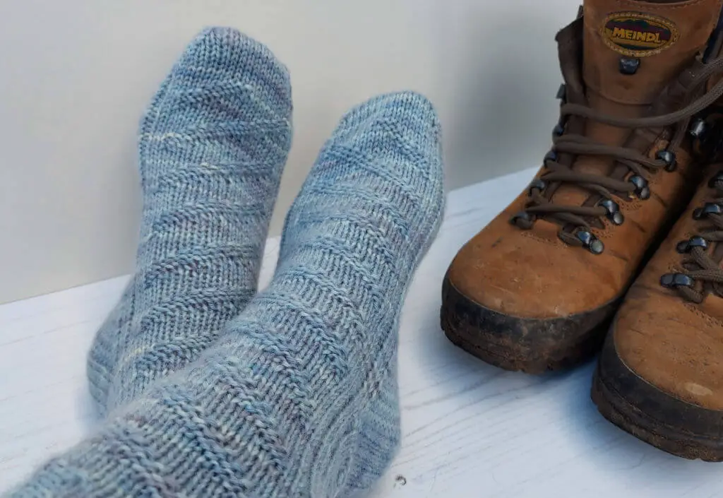 Two feet in cosy, textured blue socks rest beside a pair of worn brown hiking boots on a white wooden floor, conveying warmth and comfort.