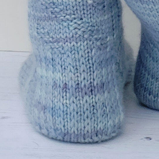 Close-up of two light blue knitted baby booties on a white wooden surface. The soft texture and pastel colour create a cosy, serene atmosphere.