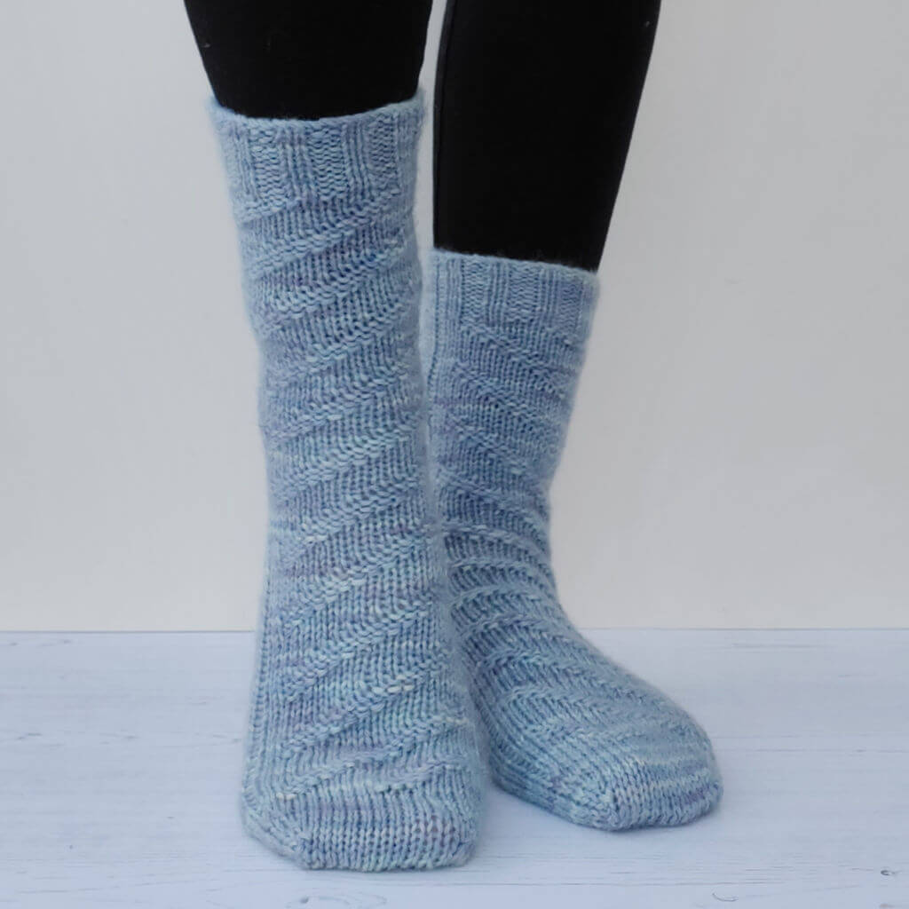 Two legs wearing light blue hand-knitted socks with a textured zigzag pattern, standing on a light wooden floor against a neutral background. Cosy and warm vibe.