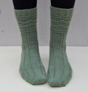 Green knitted socks on feet against a light wooden background. The socks feature a ribbed and textured pattern.