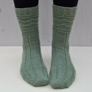 Green knitted socks on feet against a light wooden background. The socks feature a ribbed and textured pattern.