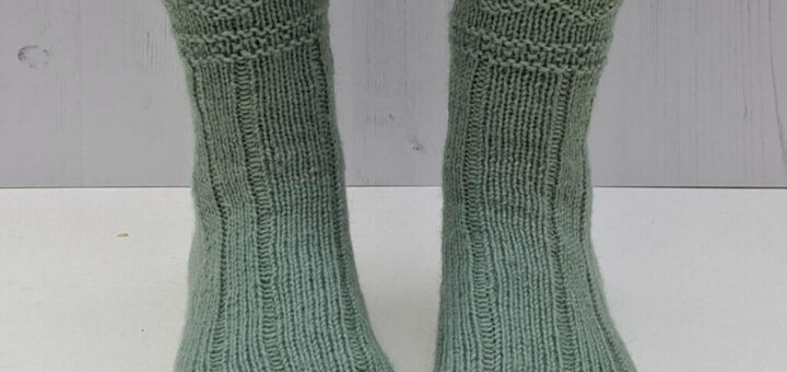 Green knitted socks on feet against a light wooden background. The socks feature a ribbed and textured pattern.