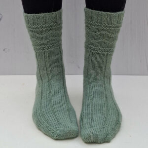 Green knitted socks on feet against a light wooden background. The socks feature a ribbed and textured pattern.