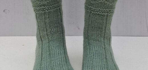 Green knitted socks on feet against a light wooden background. The socks feature a ribbed and textured pattern.